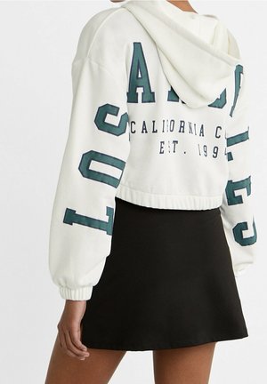 Personne portant un sweat à capuche court blanc avec des lettres vertes et une mini-jupe noire, vue de dos sur un fond uni.