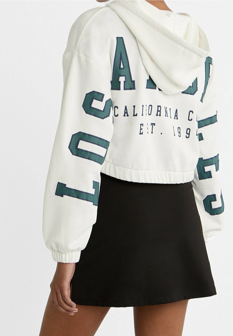Personne portant un sweat à capuche court blanc avec des lettres vertes et une mini-jupe noire, vue de dos sur un fond uni.