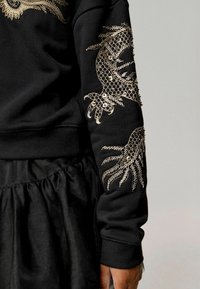 Sudadera negra con intrincados adornos dorados de diseños de peces y plantas en la manga; cuenta con puños acanalados y una textura suave.