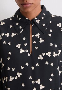 Czarna bluzka z ostrym kołnierzem i przednim dekoltem w kształcie otworu. Ozdobiona białymi wzorami motyli na całej powierzchni. Gładka struktura tkaniny.