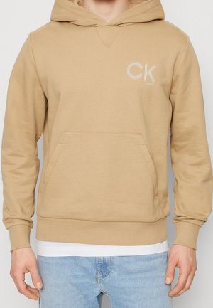 Haut à capuche en coton beige avec une poche kangourou, un capuchon réglable par cordon et un logo "CK" brodé sur la poitrine. Texture lisse, coupe décontractée.