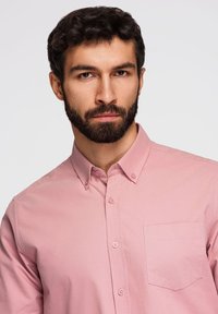 Homme aux cheveux bouclés et à la barbe portant une chemise rose boutonnée avec une poche poitrine, regardant directement l'appareil photo sur un fond uni.