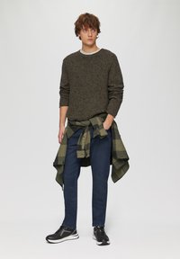Donkere gebreide trui, lichte crew neck t-shirt eronder, olijfgroene geblokte overhemd om de taille geknoopt, blauwe jeans, zwarte sportschoenen met witte zolen.
