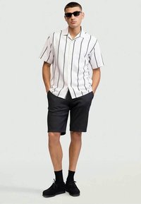 Camicia bianca a maniche corte con righe verticali nere, abbinata a pantaloni corti scuri. Il modello indossa occhiali da sole neri e scarpe nere con calzini.
