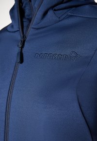 Norrøna kapuljača u tamnoplavoj boji s glatkom teksturom, s prednjim patentnim zatvaračem, podesivom kapuljačom i izvezenim logotipom na prsima.