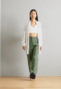 Modell som bär en vit skjorta, en kort vit topp och olivgröna byxor. Outfiten har en avslappnad passform och mjuka texturer. Svarta skor fulländar looken.
