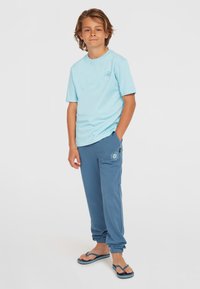 Ljusblå bomullströja och blå joggingsbyxor. T-shirten har en liten grafik, byxorna har resår i midjan och muddar. Bärs med flip-flops.