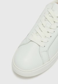 Sapatilha de couro branca com biqueira arredondada, atacadores brancos texturizados e superfície lisa com detalhes de costura mínimos.