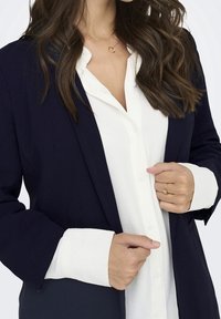 Granatowa marynarka na białej koszuli zapinanej na guziki. Marynarka ma dopasowany krój; koszula ma długie rękawy z widocznymi mankietami. Prosta złota obrączka jako dodatek.