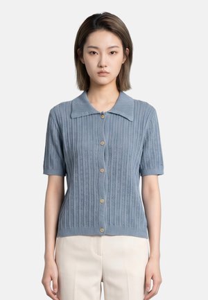 Jonge vrouw met kort bruin haar die een blauw gebreid kort mouwen gestippeld vest en lichtbeige broek draagt, naar voren kijkend.