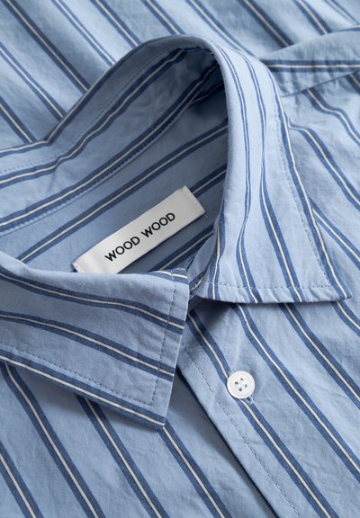 Camisa azul claro con rayas verticales en azul oscuro y blanco, cuello abierto, botón blanco y etiqueta "WOOD WOOD" dentro del cuello.