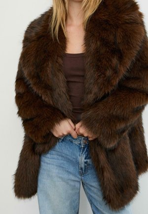 Person trägt einen dunkelbraunen Kunstpelzmantel über einem braunen Tanktop und hellblauen, hoch taillierten Jeans, Hände nahe am Mantelverschluss.