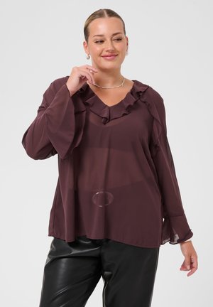 Femme aux cheveux tirés en arrière, portant un chemisier transparent violet foncé avec des volants et un pantalon en cuir noir, posant avec une main près du visage.