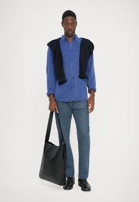 Blå skjorte med knapper, marineblå genser drapert over skuldrene, mørke jeans og svarte lærsko. Svart skulderveske.