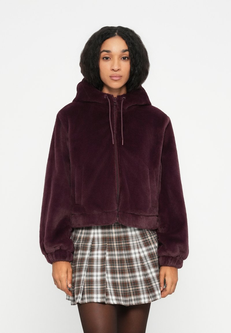 Woman wearing a dark purple plush zip-up hoodie and a brown, white, and black plaid pleated skirt with black tights.