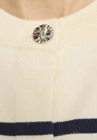 Kremkrāsas adīts džemperis ar ribotu kakla izgriezumu un izteikti spīdīgu sudraba pogu ar teksturētu virsmu, ko papildina tumši zils svītra.