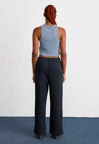 Donna con lunghe trecce arancioni, che indossa un top corto senza maniche blu, pantaloni larghi neri e scarpe con plateau nere, in piedi su un tappeto blu.