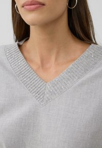 Frau, die einen hellgrauen V-Ausschnitt-Pullover mit kleinen Pailletten am gerippten Ausschnitt und silbernen Creolen trägt.