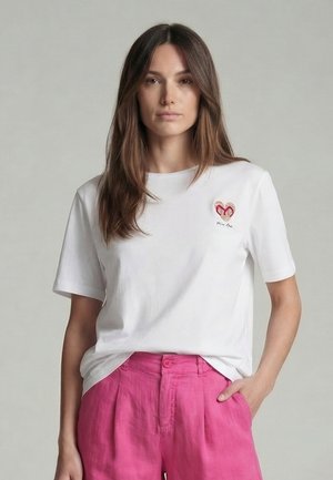 Woman with long brown hair wearing a white t-shirt with a heart patch and pink high-waisted pants, standing with one hand in pocket.