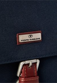 Donkerblauwe stoffen tas met een bruin leren hengsel, voorzien van een zilveren metalen label met de tekst "TOM TAILOR" en een metalen gesp.