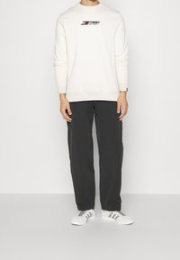 Felpa crema con texture a coste e logo Tommy Hilfiger, abbinata a pantaloni cargo scuri e sneakers bianche con strisce grigie.