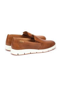 Zapatos tipo mocasín de cuero marrón con un diseño texturizado, banda elástica y suela de goma blanca, con una discreta etiqueta de marca en el lateral.