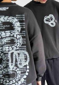 Schwarzer Pullover mit großem weißen Schlangendesign und Text, der verschiedene Städte auf dem Rücken auflistet. Gerippter Kragen und Bündchen.
