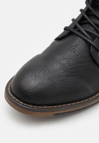 Černá kožená bota s kulatou špičkou, texturovaným povrchem a hnědou gumovou podrážkou. Obsahuje šněrovací design a kontrastní prošívání na okraji.