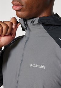Grå og sort Columbia vindjakke med lynlås foran, justerbar hætte med snoretræk og fremtrædende logo på brystet.