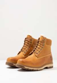 Braune Lederstiefeletten mit synthetischem Fellfutter, metallischen Ösen und einer Gummisohle mit horizontalen Profilmustern. Timberland-Logo an der Seite.