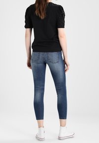 Mujer con una blusa negra de manga corta con mangas de cuentas, jeans ajustados cortos azules y zapatillas altas blancas, de pie de espaldas.