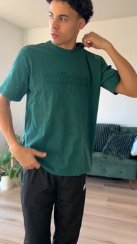 Žalia medvilninė marškinėliai su reljefiniu „adidas“ logotipu, apvali iškirptė ir trumpomis rankovėmis, derinami su juodomis kelnėmis, ryškioje vidaus aplinkoje.