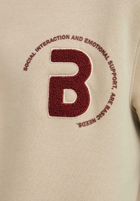 Sudadera beige con un logo "B" testurizzato in mogano. Il testo circostante recita: "L'INTERAZIONE SOCIALE E IL SUPPORTO EMOTIVO SONO BISOGNI FONDAMENTALI."