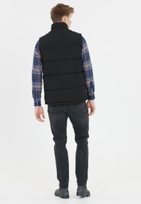 Schwarze Steppweste mit hohem Kragen, horizontalen Nähten und einer glatten Stoffstruktur. Kombiniert mit einem blauen Karoshirt und dunkler Jeans.
