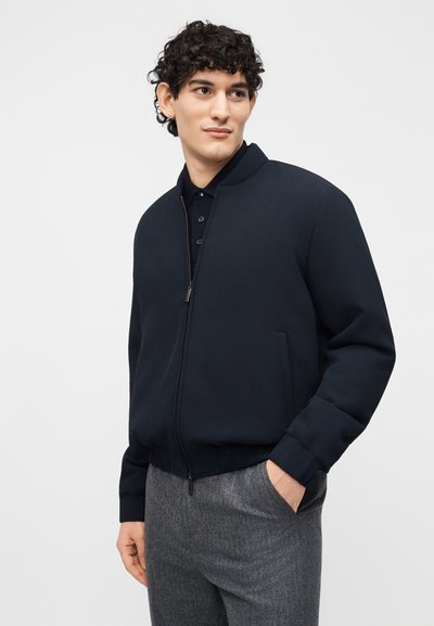 Νεαρός με κατσαρά μαλλιά, φοράει μπλε μπουφάν με φερμουάρ πάνω από μαύρο πόλο και γκρι παντελόνι, στέκεται με το ένα χέρι στην τσέπη.