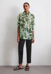 Chemise verte en voile à boutons avec motif floral, portée avec un pantalon droit noir et des sandales noires avec une boucle dorée.