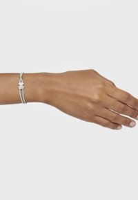 Pulsera ajustable con dos cuerdas de color azul claro y un charm en forma de estrella blanca. Lleva puesta en una mano bronceada, con una textura suave.