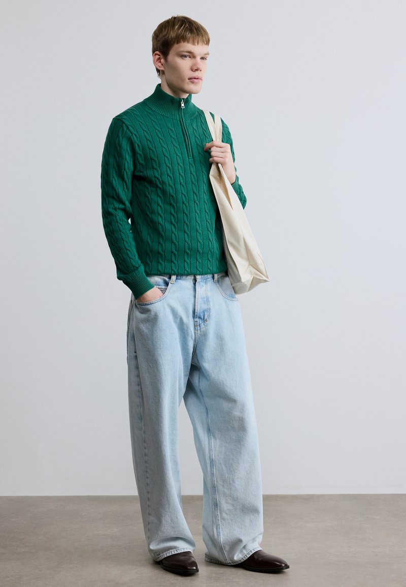 Green cable-knit sweater with high collar, paired with loose light blue denim jeans and black pointed shoes. Model holds a cream tote bag.