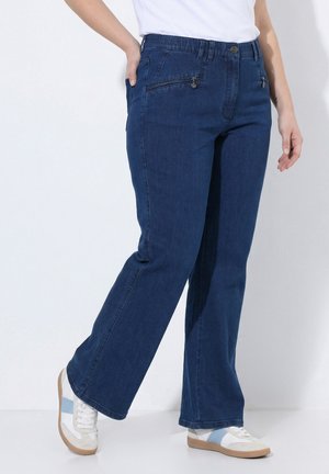 Vrouw draagt donkerblauwe jeans met rechte pijpen en witte sneakers met blauwe strepen, staat met één hand in de zak.