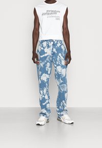 Valkoinen hihaton toppi, jossa painettu teksti, yhdistetty vaaleansinisiin farkkuihin, joissa graafinen kuvio. Valkoiset tennarit täydentävät asun.