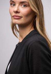 Černá bunda na zip s texturovaným límcem, s hladkým povrchem látky a přiléhavým střihem. Detailní záběr na profil modelky.