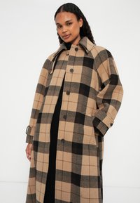 Manteau à carreaux en tan et noir, fabriqué en mélange de laine. Comprend un col, de grands boutons et des poches latérales. Coupe légèrement oversize avec des manches longues.