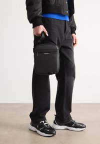 Borsa nera con texture, dotata di manico superiore e chiusura a zip, portata da una persona in pantaloni scuri e sneakers nere. Design minimalista.