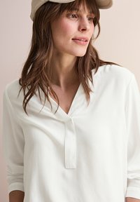 Haut à manches longues blanc avec un décolleté en V et un tissu texturé. Détail de bouton notable au niveau du cou. Porté avec une casquette de couleur claire.