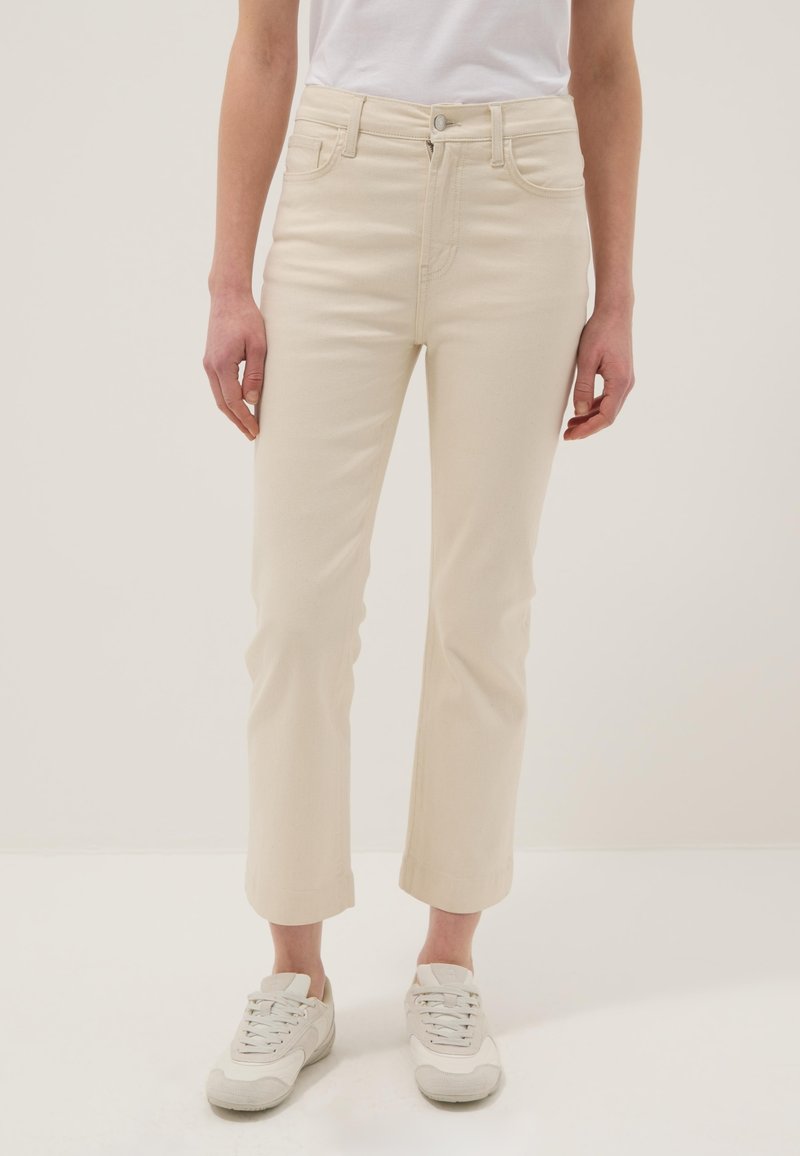 Persona vistiendo pantalones rectos color hueso y zapatillas blancas, de pie contra un fondo claro y liso.