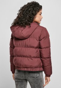 Giacca a vento bordeaux con cappuccio, caratterizzata da un design trapuntato e polsini elasticizzati. Il materiale appare liscio e leggermente lucido.