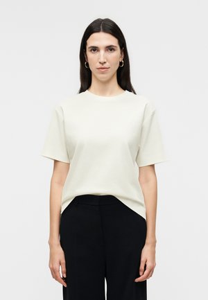 Γυναίκα με ίσια σκούρα μαλλιά, φορώντας χρυσούς κρίκους στα αυτιά, κοντομάνικη εκρού μπλούζα και μαύρο παντελόνι, σε απλό λευκό φόντο.