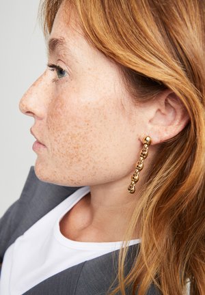 Pendientes de cadena de oro con elementos esféricos unidos, reflejando la luz. La modelo lleva una chaqueta de traje gris sobre una camisa blanca, vista de perfil lateral.