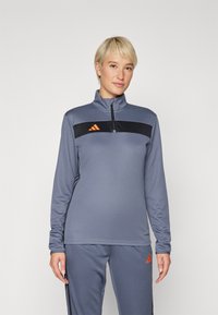 Sivi sportski top s polu-zip ovratnikom, crnom prugom preko prsa i narančastim Adidas logom. Glatka tekstura i prilagođeni dizajn.