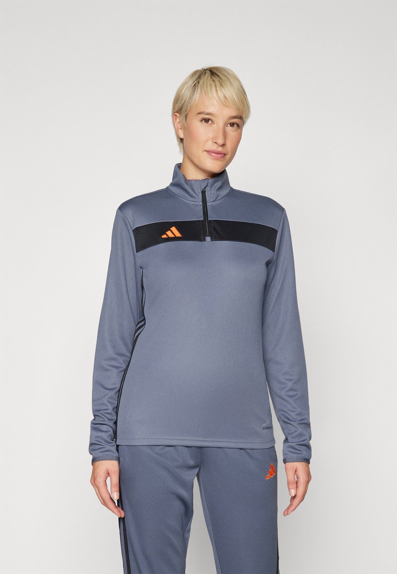 Sivi sportski top s polu-zip ovratnikom, crnom prugom preko prsa i narančastim Adidas logom. Glatka tekstura i prilagođeni dizajn.